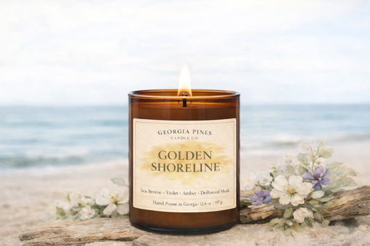 Candle labeled 'Golden Shoreline' on a beach with flowers and driftwood.